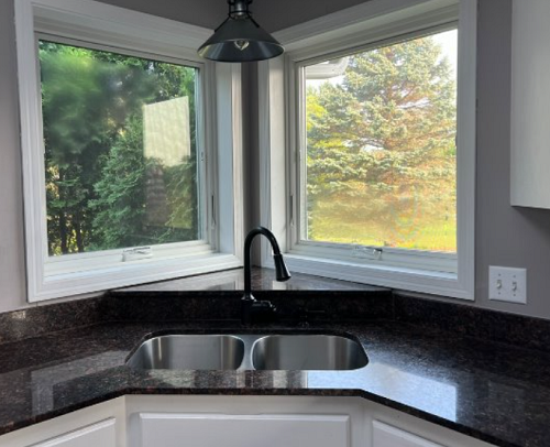 Kitchen corner with double sink below two white‑framed windows showing outdoor trees