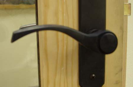 Close-up of a new oil rubbed bronze door handle that is all black and mounted on a black rectangular backplate, attached to a light-colored wooden door frame next to a glass panel.