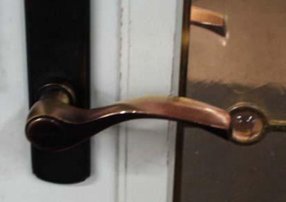 Close-up of a curved, seasoned bronze lever door handle on a rectangular black mounting plate, attached to a white door frame next to a glass panel.