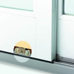 Close-up of the bottom corner of a white sliding door or window frame on a metal track. A circular cutout in the frame reveals two metal roller wheels used to guide the door along the track.
